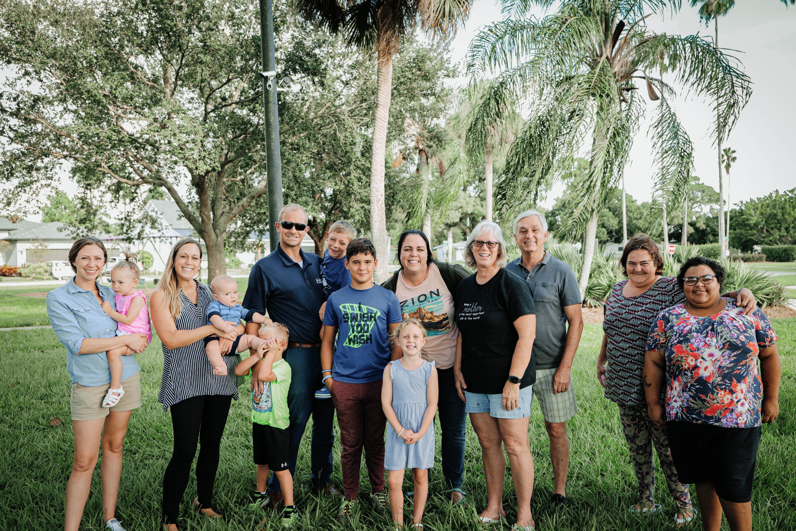 14 people gather for a group photo on a span of green grass surrounded by palm trees